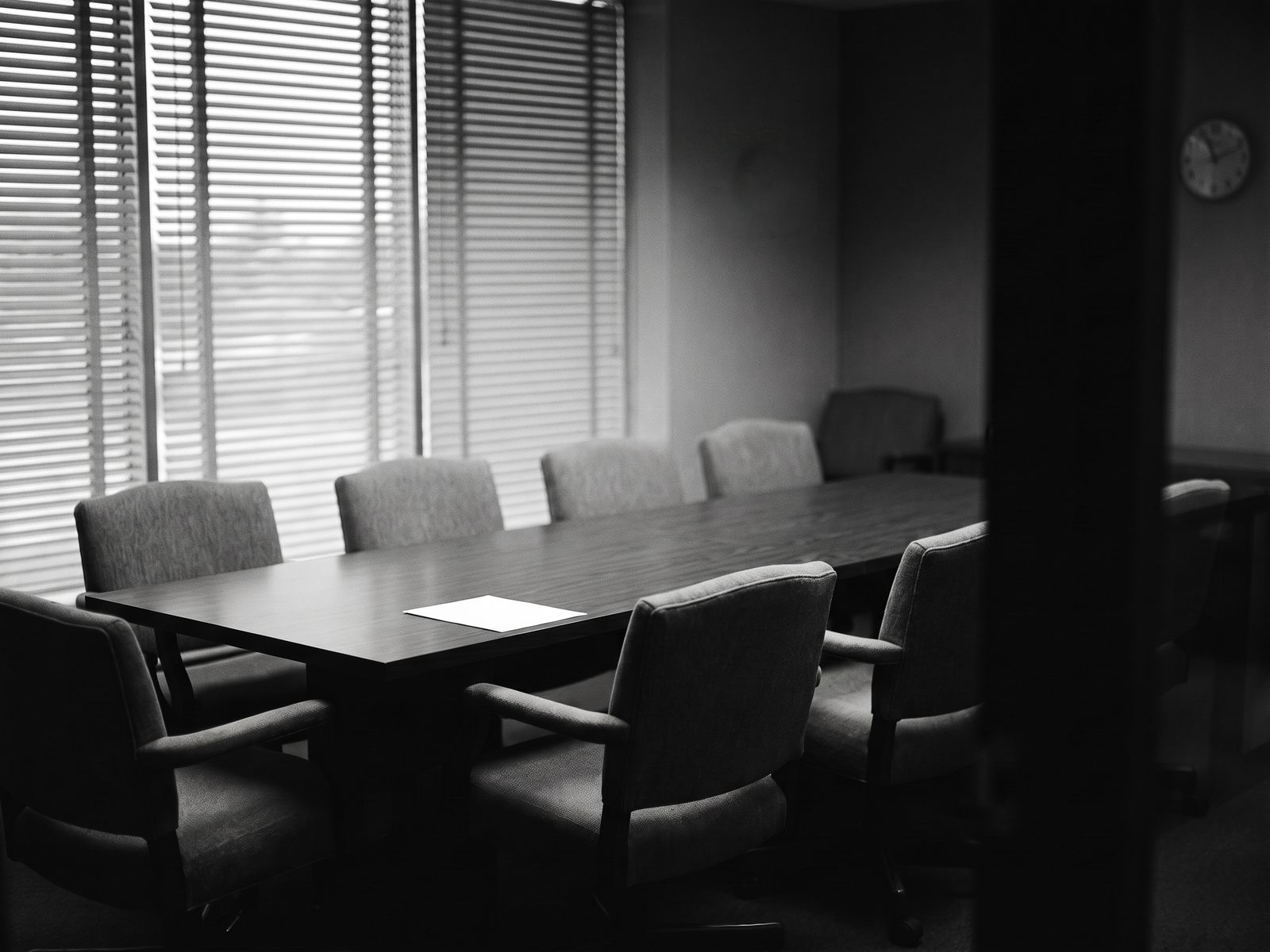 An empty conference room after hours: eight chairs around a long table, one sheet of paper, a wall clock.